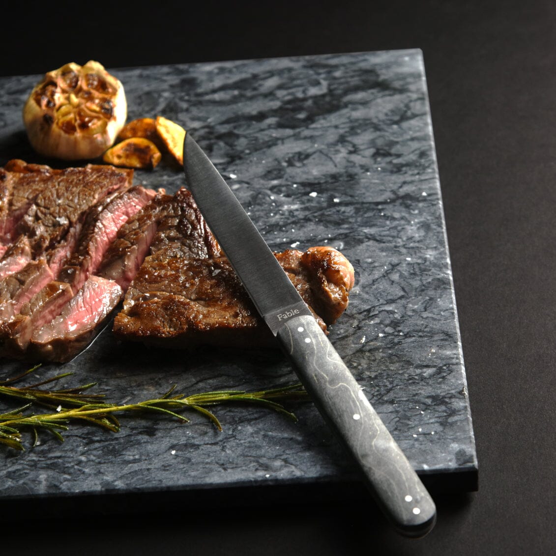 Sliced steak on a dark marble slab with a knife, garlic, and rosemary.